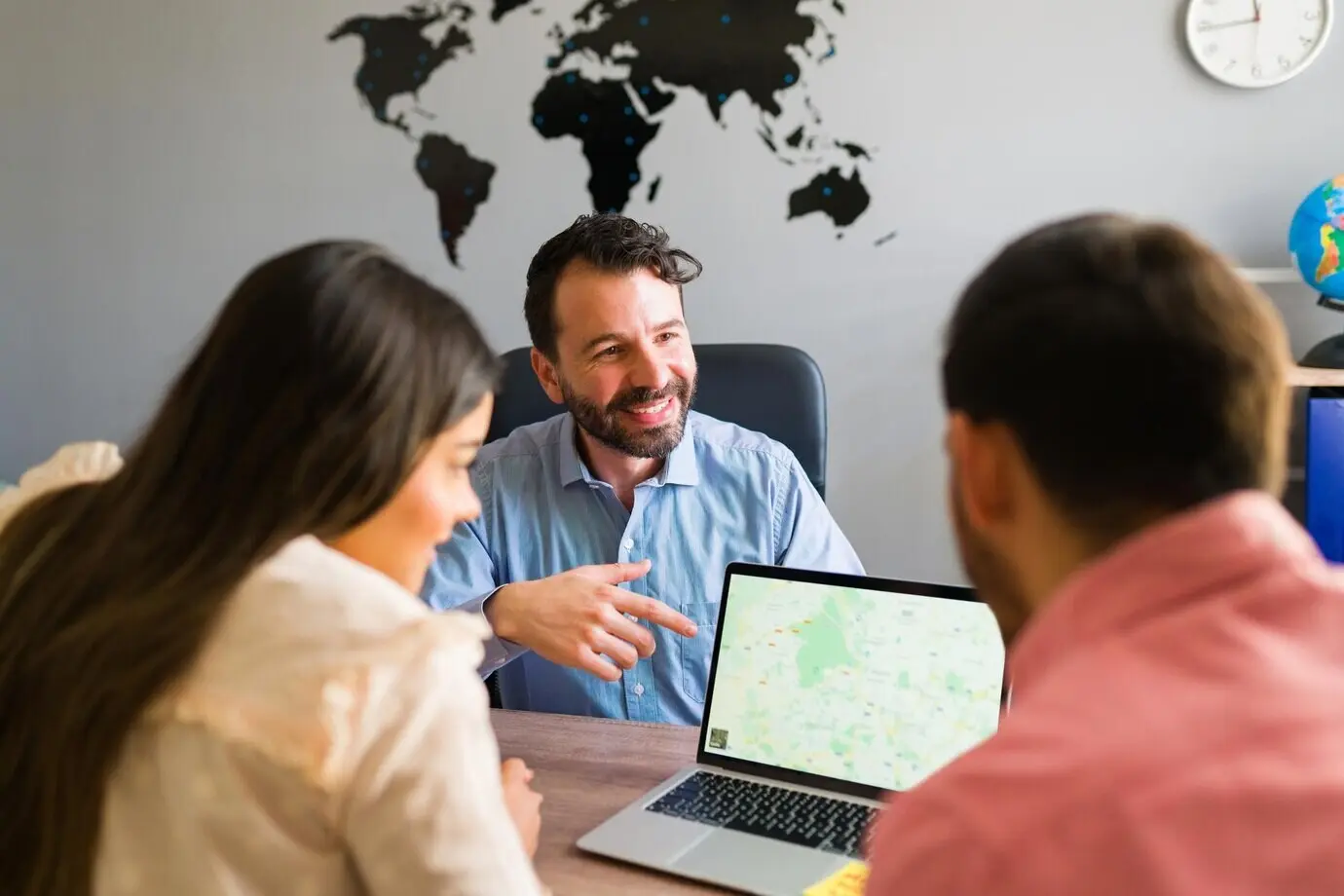 Your next trip destination awaits. Seen from behind, a beautiful couple at a travel agency looks at a map on a laptop, choosing where to go on vacation.